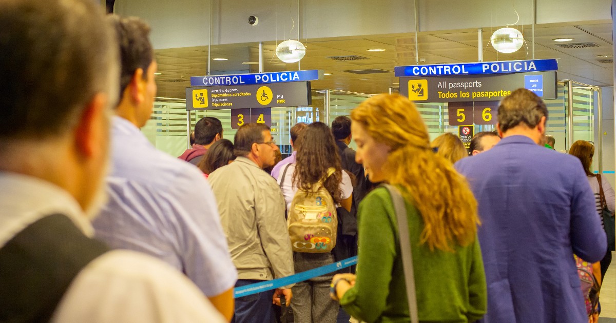Imaginea prezintă o coadă de pasageri care așteaptă la ghișeele de control al pașapoartelor dintr-un aeroport.