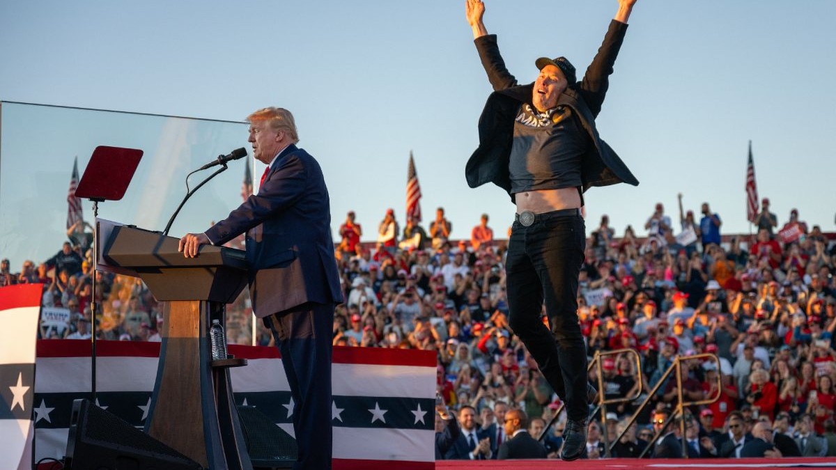 Donald Trump, pe o scenă, în timpul campaniei electorale, îmbrăcat în costum, iar alături de el, Elon Musk, îmbrăcat cu blugi, tricou și sacou, în timp ce sare în sus, cu mâinile îndreptate spre cer