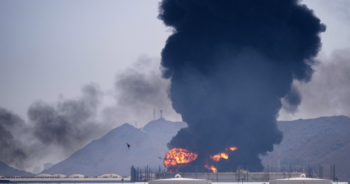 Imaginea surprinde o explozie violentă la o instalație industrială, cel mai probabil o rafinărie sau un depozit de combustibil, cu flăcări și o coloană masivă de fum negru care se ridică spre cer.