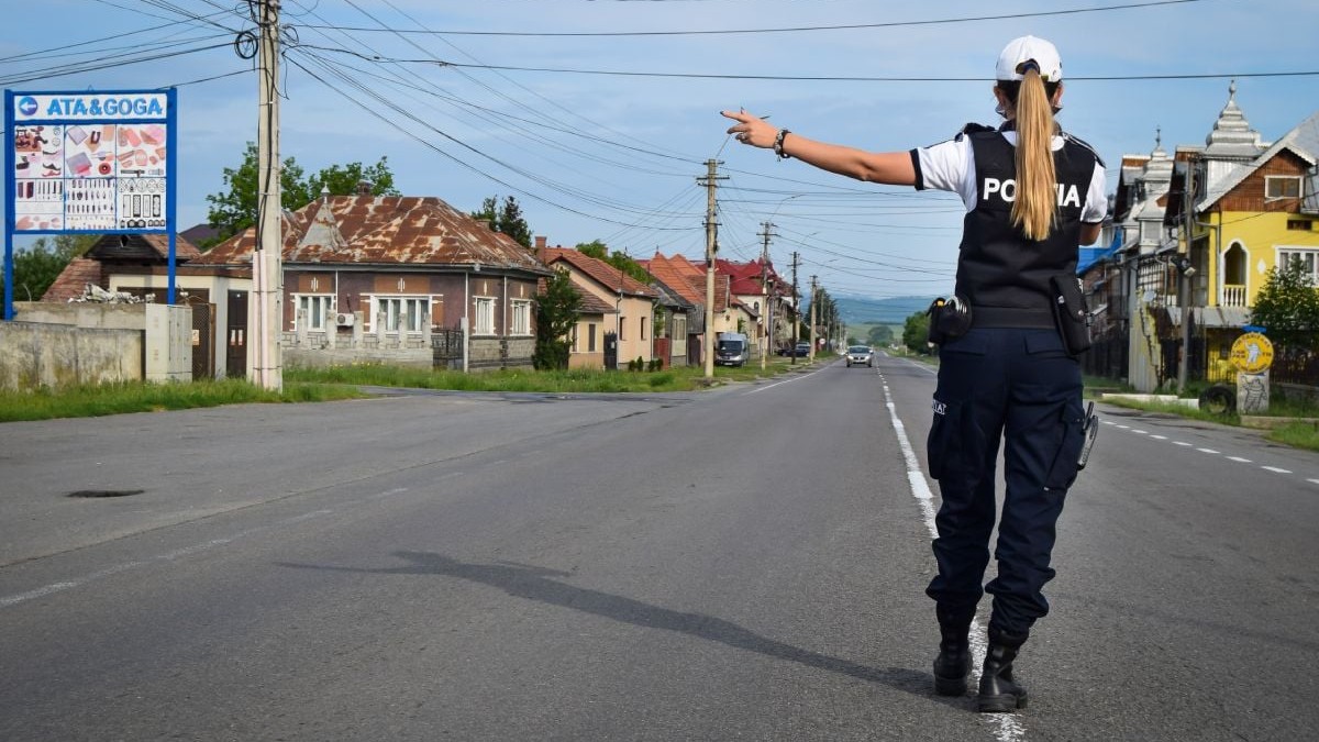 Polițistă de la Rutieră, îmbrăcată în uniformă, cu șapcă albă, pe mijlocul unei străzi din Cluj-Napoca, fotografiată din spate, cu mâna stângă întinsă, în timp ce oprește un șofer