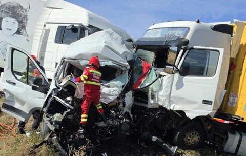 Imaginea prezintă urmările unui accident grav, în care o dubă albă este zdrobită de două TIR-uri, în timp ce un echipaj de pompieri și paramedici SMURD intervine la fața locului.
