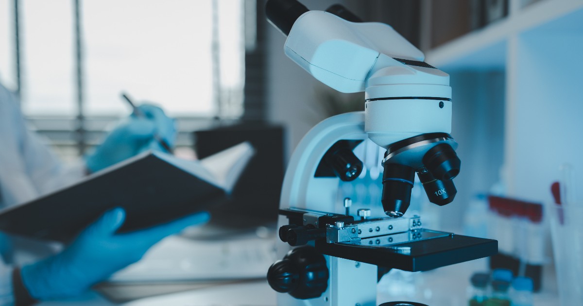 Imaginea arată un microscop de laborator utilizat într-un mediu de cercetare biomedicală, cu detalii clare asupra instrumentului. În fundal, un cercetător notează observații într-un caiet, sugerând desfășurarea unui experiment științific.