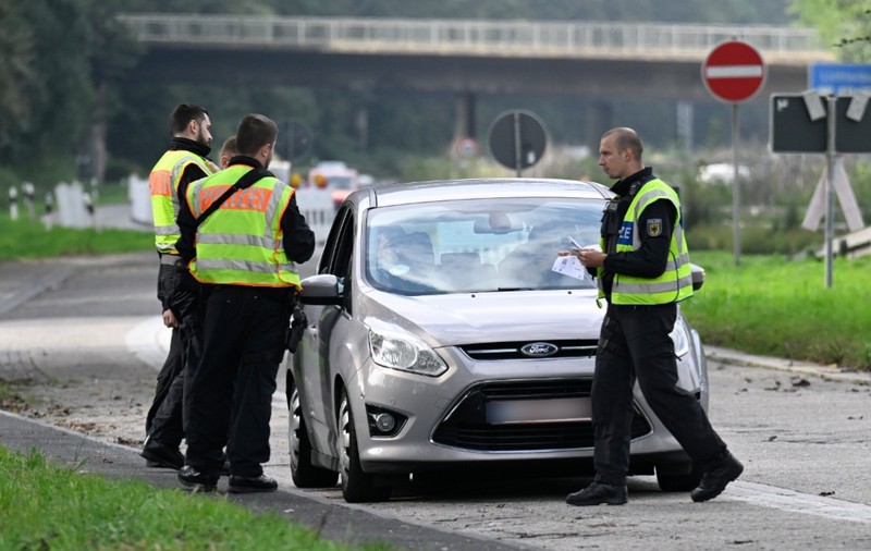 Polițiști germani purtând veste reflectorizante care efectuează un control de rutină asupra unui autoturism Ford argintiu oprit la marginea unui drum.