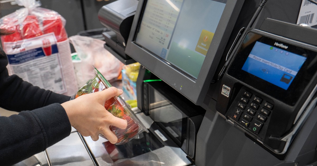 Imaginea surprinde un client care scanează un produs la o casă de marcat self-pay dintr-un supermarket. Fotografia ilustrează procesul de autoservire și utilizarea tehnologiei moderne în comerțul de retail.
