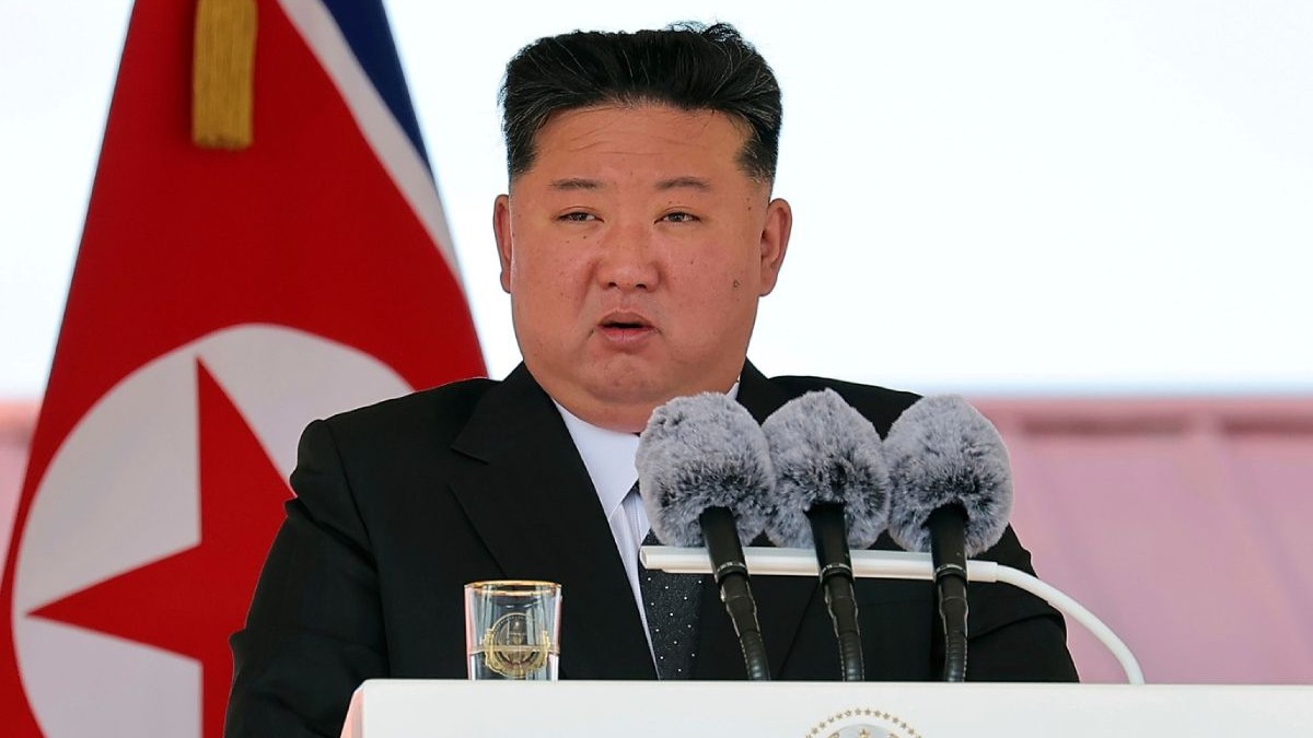 Liderul Nord Coreean Kim Jong Un, in sacou negru si camasa albă, in timp ce vorbește la un pupitru alb, cu trei microfoane pe el si un pahar alături, in cadrul unei conferinte, 12 iunie 2025, șantierul naval Rajin din Rason, Coreea de Nord