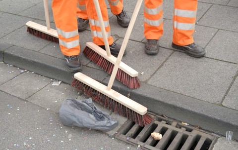Picioarele unor lucrători de la salubritate purtând pantaloni portocalii reflectorizanți și măturând un trotuar și bordura unei străzi acoperite de resturi și deșeuri.   