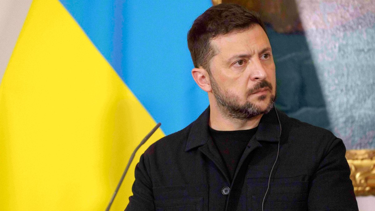 Imaginea prezintă un portret de prim-plan al unui bărbat cu barbă scurtă, îmbrăcat într-o jachetă neagră cu nasturi, având o expresie serioasă și concentrată. În fundalul din stânga, se poate observa clar steagul galben și albastru al Ucrainei.