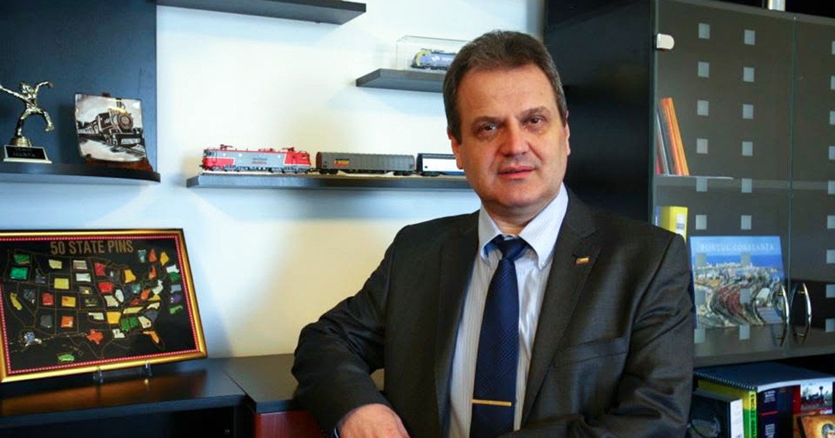 George Micu îmbrăcat într-un costum închis la culoare stă rezemat de o piesa de mobilier.