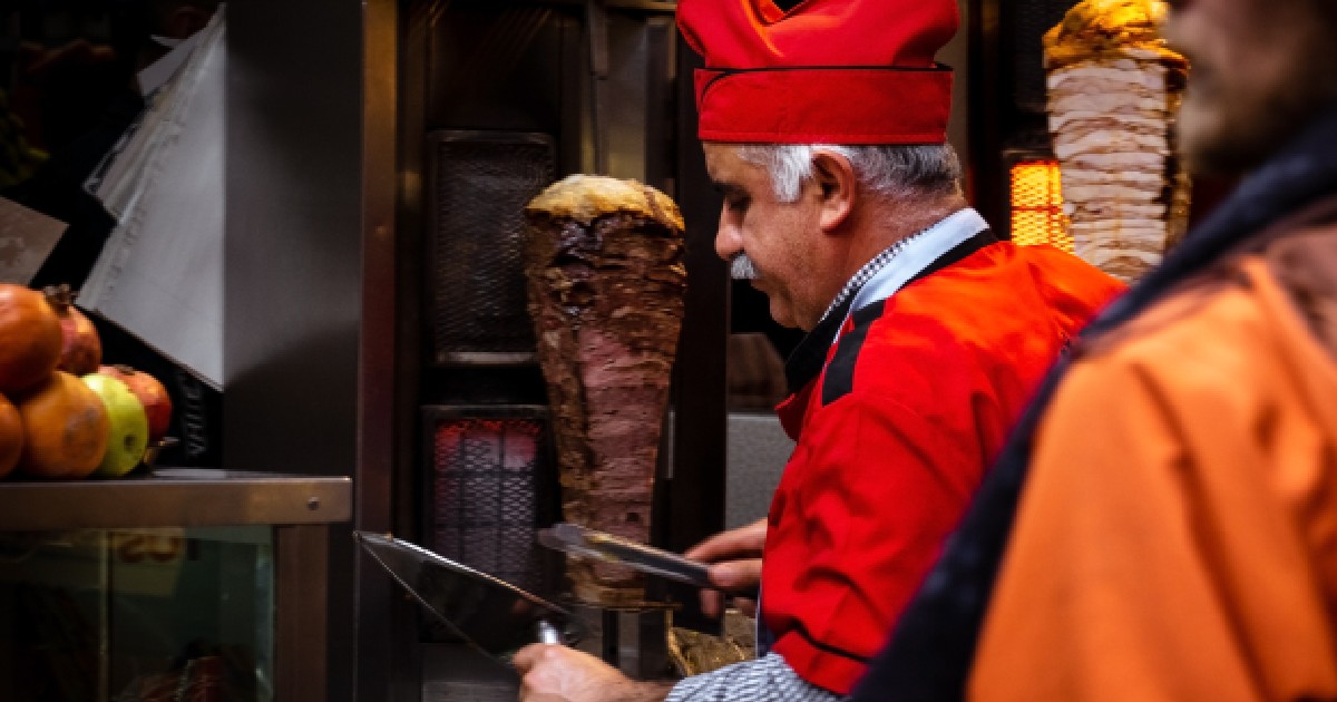 Imaginea surprinde un bucătar îmbrăcat într-o uniformă roșie cu o căciulă roșie specifică, în timp ce pregătește un doner kebab tăind straturi subțiri de carne de pe un frigărui vertical rotativ. Atmosfera este întunecată și pune în evidență carnea gătită și lumina roșiatică a elementelor de încălzire.