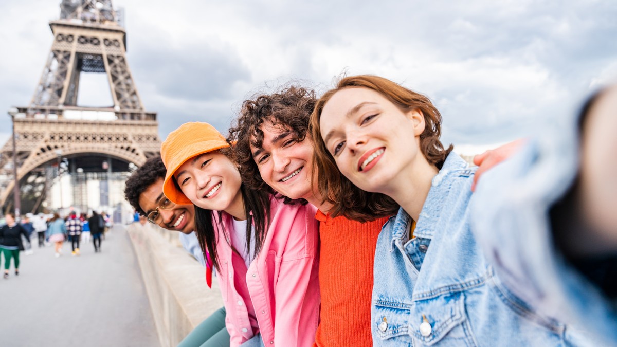 grup de tineri turiști la Paris își fac un selfie cu Arcul de Triumf pe fundal, zâmbind fericiți