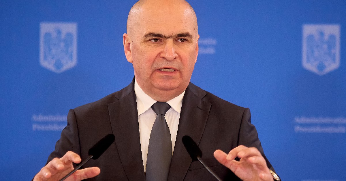 Imaginea îl înfățișează pe Ilie Bolojan susținând o declarație de la o tribună, gesticulând cu ambele mâini în fața unui fundal albastru ce poartă însemnele Administrației Prezidențiale.