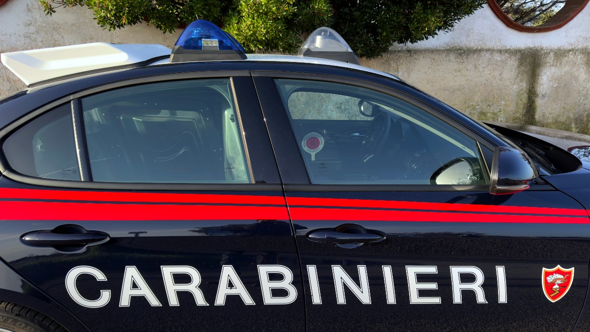Imaginea prezintă partea laterală a unei mașini de patrulă de culoare închisă (negru/bleumarin), aparținând forței de poliție italiene, Carabinieri. Mașina are inscripția "CARABINIERI" cu litere mari albe și o bandă orizontală lată de culoare roșie, completată de girofaruri albastre pe plafon.