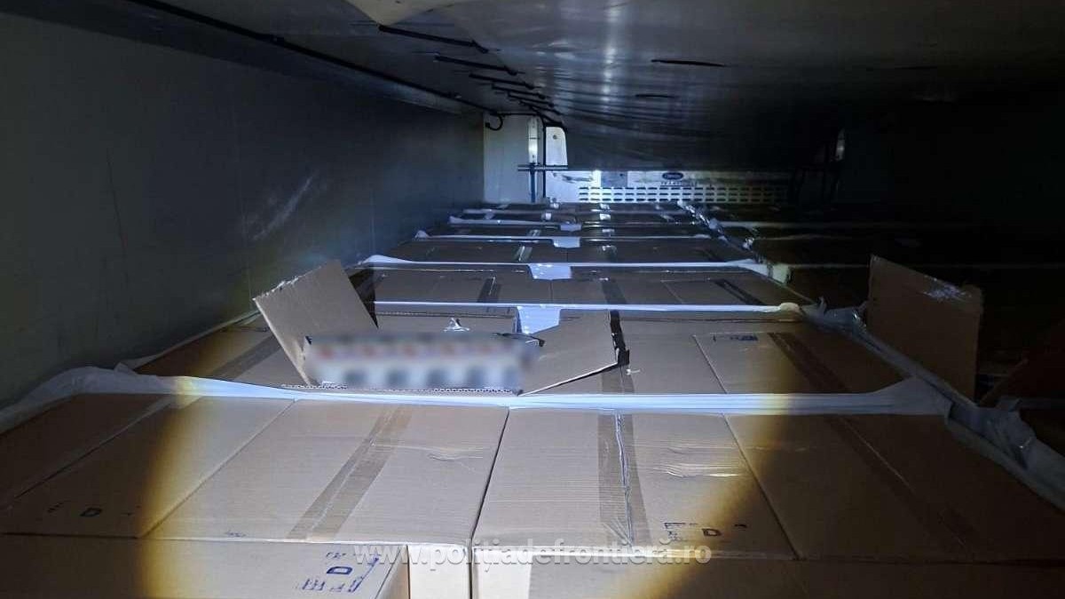 Imaginea arată interiorul unui spațiu restrâns, probabil o remorcă de camion sau un container, umplut de jos în sus cu numeroase cutii de carton mari, ambalate. Un spațiu îngust este lăsat liber, permițând vizualizarea plafonului metalic și a rândurilor de cutii care se extind în adâncime.