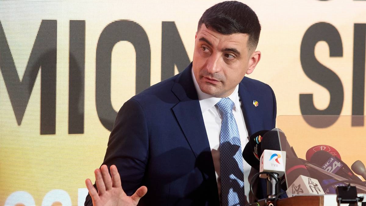 George Simion, îmbrăcat într-un costum bleumarin, cămașă albă și cravată albastră, în timpul unei conferințe de presă.