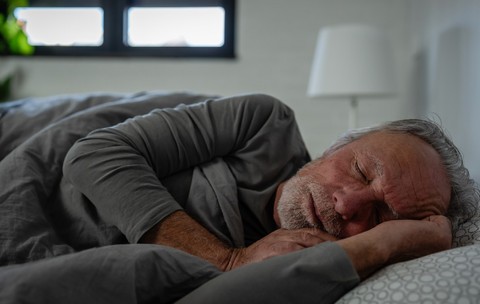 Un bărbat în vârstă care doarme liniștit în pat, întins pe o parte sub o pătură gri, într-un dormitor cu lumină difuză