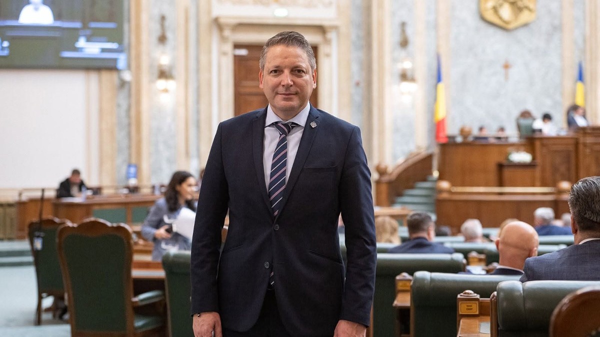 Marius Hudelmicu îmbrăcat într-un costum închis la culoare, cămașă bleu și cravată în dungi.