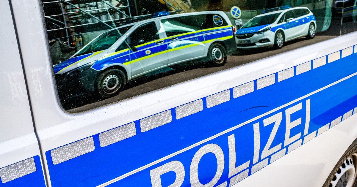 Imaginea este un prim-plan al caroseriei albe a unei autospeciale a poliției germane, marcată cu dunga lată albastră și inscripția mare "POLIZEI" în partea inferioară, alături de o serie de benzi reflectorizante gri. Pe geamul lateral se reflectă un alt microbuz de poliție și o mașină de patrulă (break) din aceeași flotă, ambele având schema de culori alb-albastru-galben, tipică forțelor de ordine din Germania.