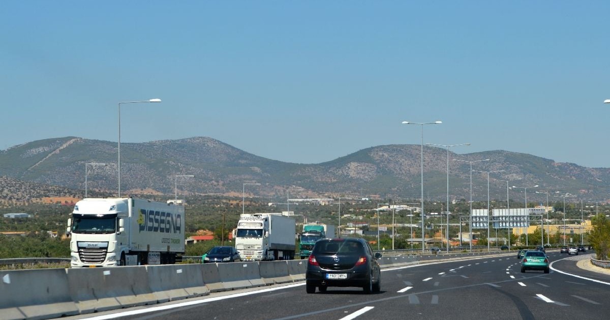 Imaginea prezintă traficul pe o autostradă cu mai multe benzi, unde circulă camioane de mare tonaj și autoturisme, pe fundalul unor dealuri, sub un cer senin.