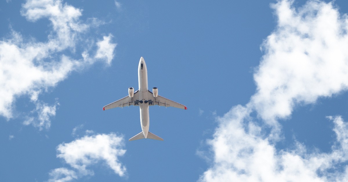 Imaginea arată un avion de pasageri alb, surprins de jos, care zboară pe un cer albastru intens presărat cu nori albi și pufoși. Avionul, care pare să fie la o altitudine joasă, are vizibil trenul de aterizare desfășurat.