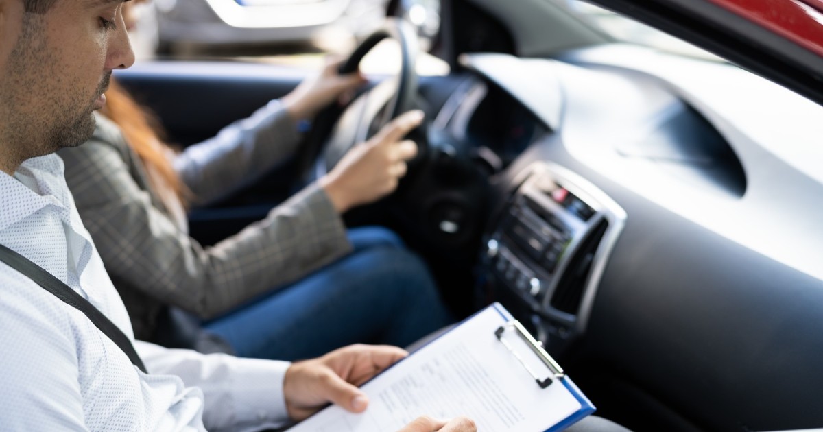În imagine este ilustrat momentul examenului pentru permisul auto. O tânără se află la volan, iar examinatorul în partea dreaptă.