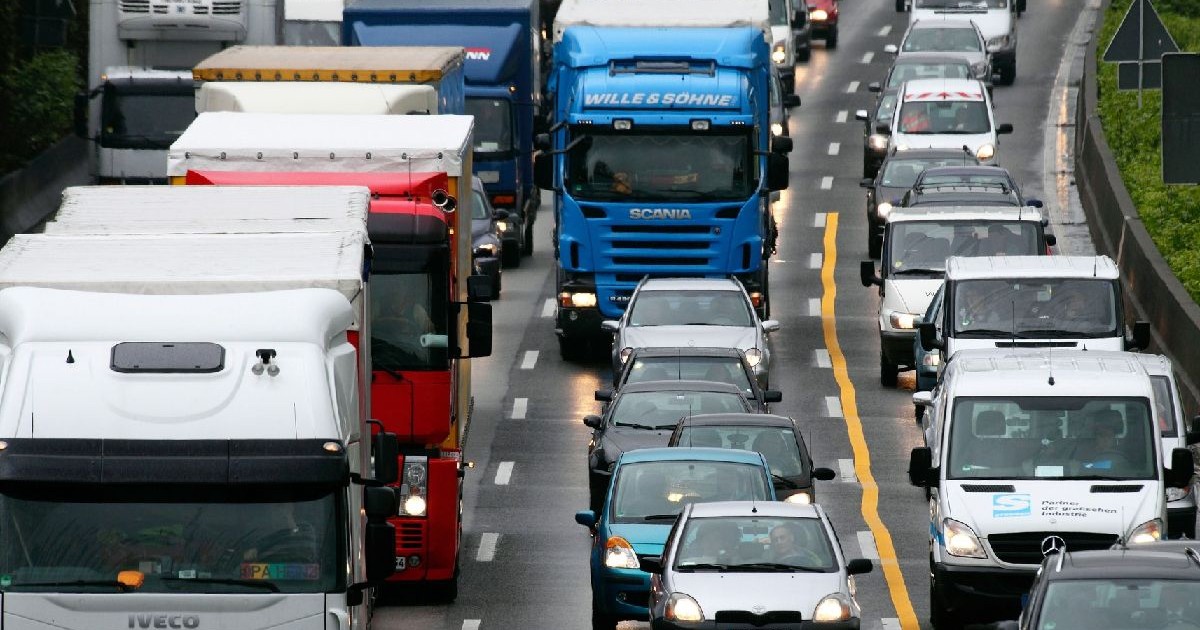 Imaginea prezintă o autostradă aglomerată, pe care circulă diverse vehicule, inclusiv camioane