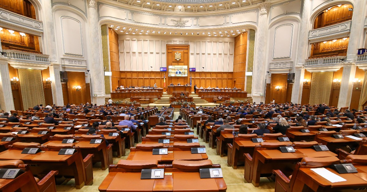 Imaginea surprinde interiorul unei săli de plen impunătoare, plină de deputați sau senatori așezați la pupitre individuale din lemn, orientate către prezidiu, sugerând o ședință a Parlamentului. Sala este caracterizată de o arhitectură neoclasică, cu pereți înalți, decorațiuni aurite și o tribună centrală mare, deasupra căreia se află Stema României.
