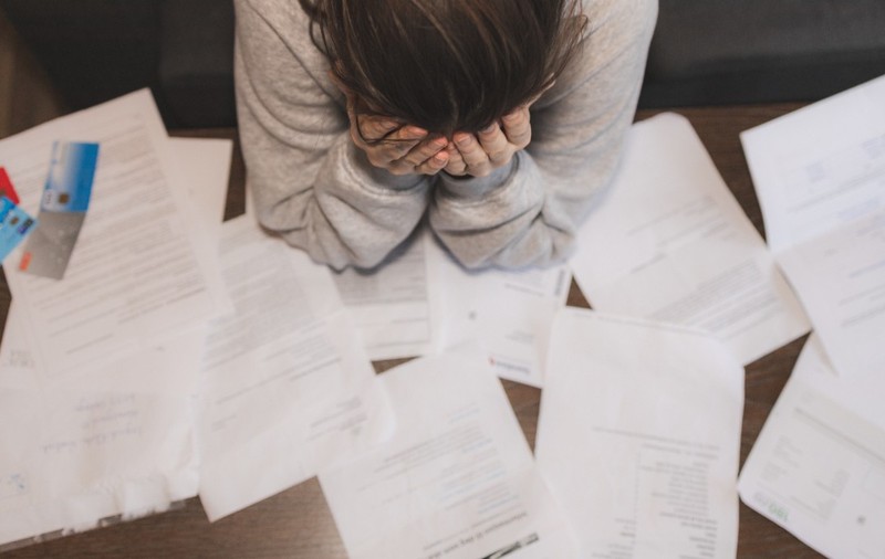 O persoană care își ține capul în mâini deasupra unei mese pline de facturi, documente împrăștiate și carduri bancare, sugerând o stare de stres sau epuizare financiară