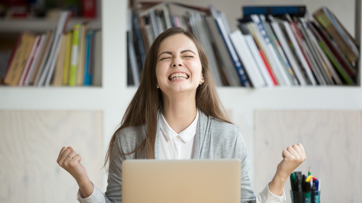 Studentă care stă la birou în fața unui laptop și râde fericită