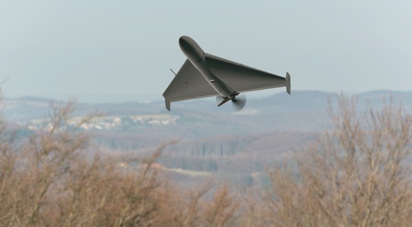O dronă militară gri în formă de aripă triunghiulară zboară la joasă altitudine deasupra unei zone împădurite și a unui peisaj deluros. Poză cu caracter ilustrativ. Foto: Shutterstock
