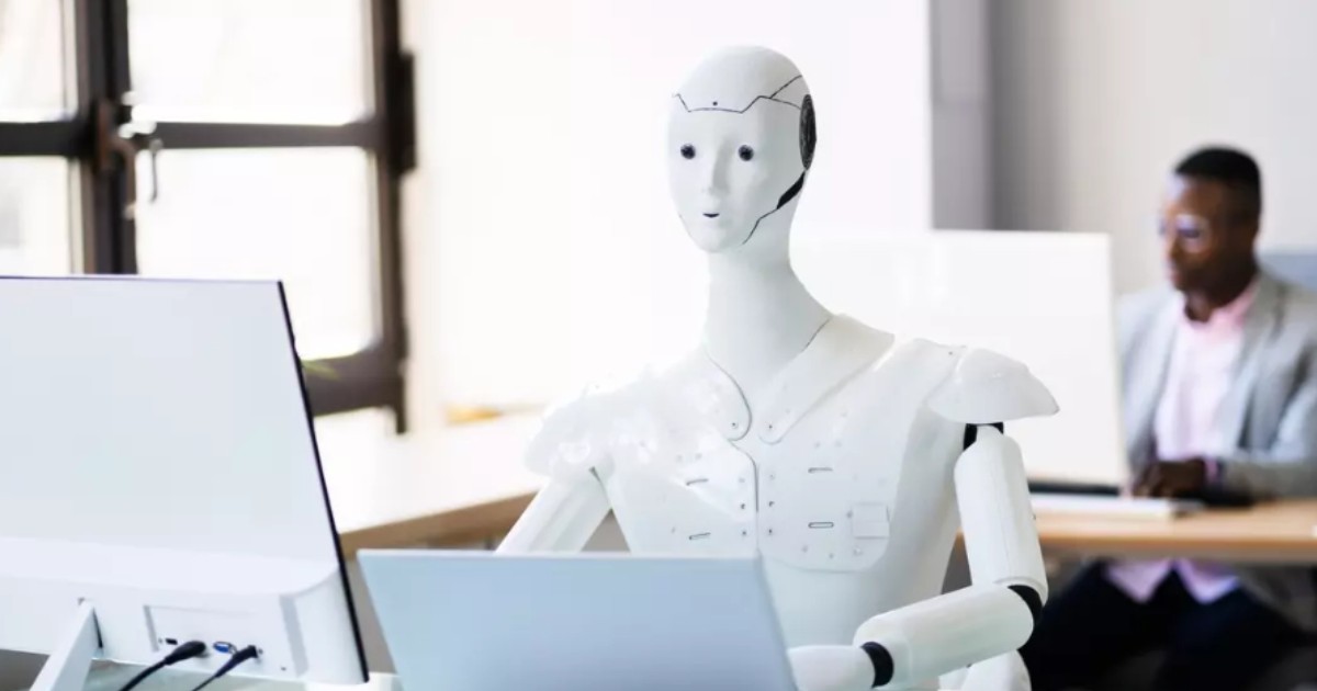 Un robot alb așezat la un birou, lucrând la un laptop.