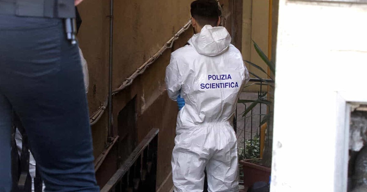 Fotografie cu un bărbat îmbrăcat într-un costum alb de protecție, cu inscripția „POLIZIA SCIENTIFICA” pe spate, examinând locul unei infracțiuni într-o încăpere îngustă și slab iluminată, cu o plantă în ghiveci și o balustradă metalică