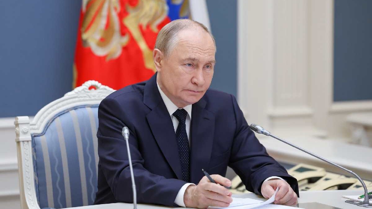Vladimir Putin în biroul său, îmbrăcat într-un sacou negru, o cămașă albă și o cravată neagră, stând cu mâinile pe masă, ținând într-una din mâini un document iar în cealaltă un pix negru