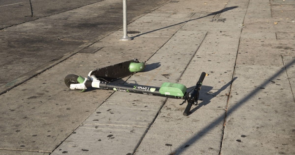O trotinetă electrică verde/neagră zace abandonată pe un trotuar de beton dintr-un oraș.