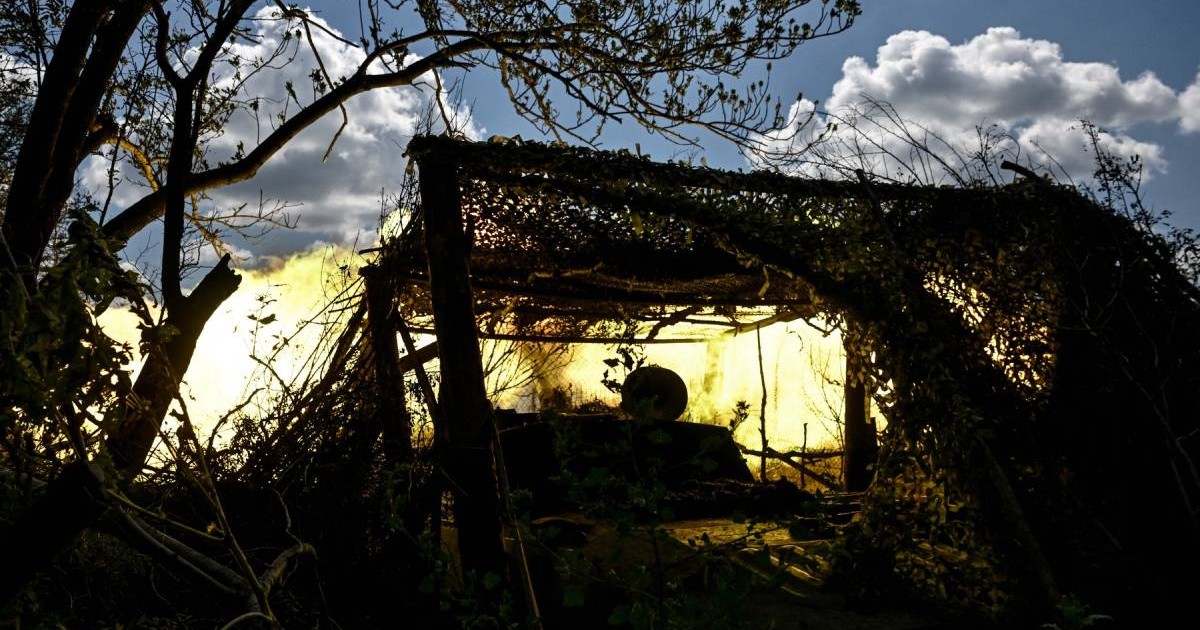 O poziție de artilerie camuflată în vegetație, luminată puternic de o explozie galben-aurie în fundal, cu crengi și frunziș care ascund silueta echipamentului militar în Pokrovsk - Ucraina