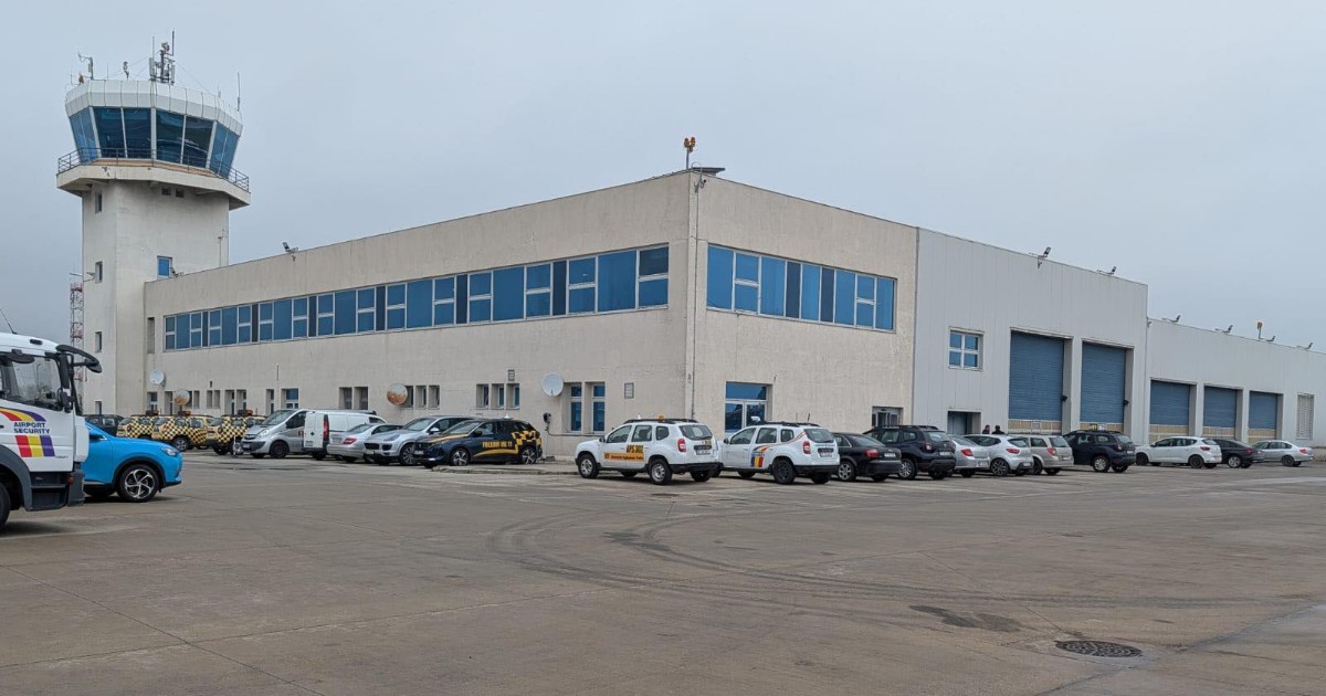Clădirea unui hangar sau centru tehnic din cadrul aeroportului este surprinsă alături de turnul de control, într-o zonă largă de platformă. În față sunt parcate mai multe mașini și vehicule de serviciu, sugerând activitatea operațională din incinta aeroportului.