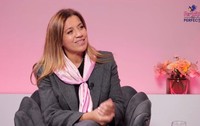 Mirela Retegan, îmbrăcată într-un costum gri, cu o eșarfă roz la gât, pe un fotoliu gri, iar pe fundal un perete roz și o vază cu flori