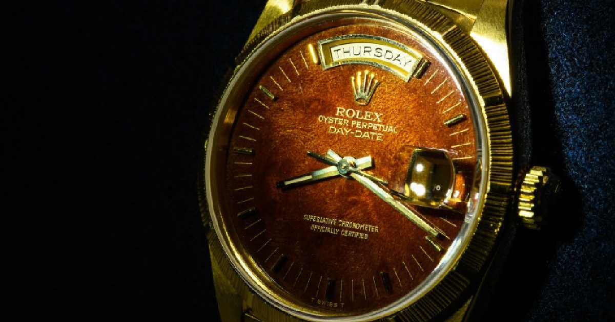 Prim-plan a unui ceas Rolex Oyster Perpetual Day-Date cu cadran maro, carcasă și brățară din aur, pe un fundal texturat de culoare albastru închis.