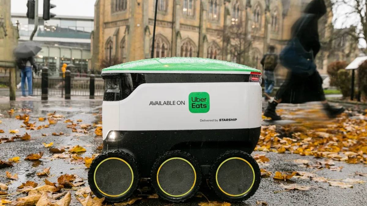 Un robot de livrare Uber Eats, de culoare alb cu verde, pe o strada tomnatică din Marea Britanie, cu frunze în jur.