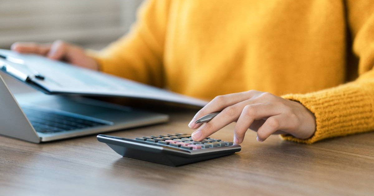 O femeie care stă în fața calculatorului, ține o serie de hârtii în mână și face calcule pe un mic calculator manual.