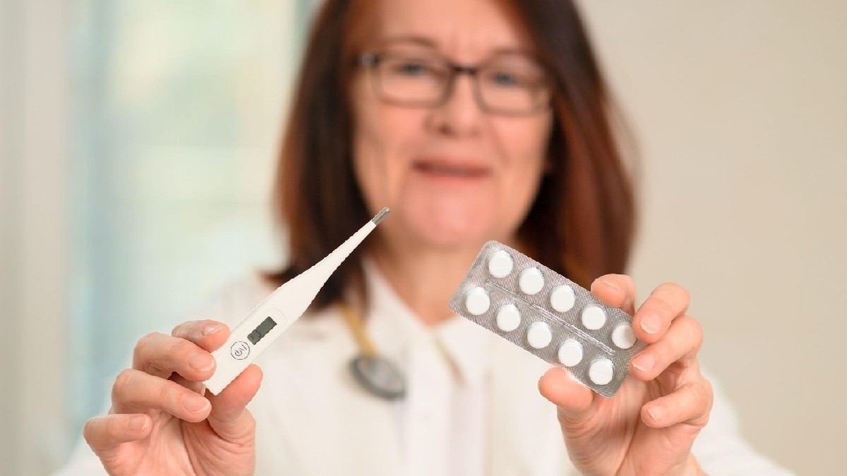 O doctoriță în uniformă albă cu stetoscop se uită la cameră. O mână ține un termometru, cealaltă pastile. Accent pe îngrijirea sănătății, verificarea febrei și