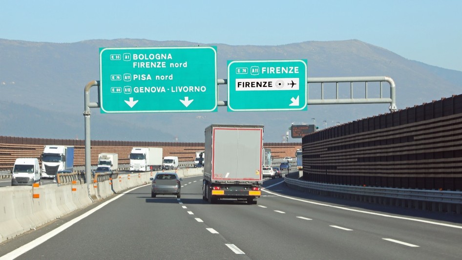 Traficul de pe o autostradă din Italia, cu mai multe autovehicule și camioane deplasându-se pe benzi sub panouri verzi mari de semnalizare rutieră ce indică direcțiile spre Bologna, Firenze, Pisa, Genova și Livorno.