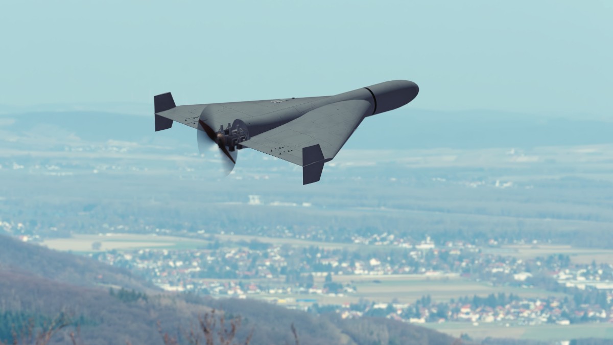 Imaginea prezintă o dronă aeriană de tip kamikaze care zboară la joasă altitudine deasupra unei zone locuite, având în fundal un peisaj cu dealuri și localități.