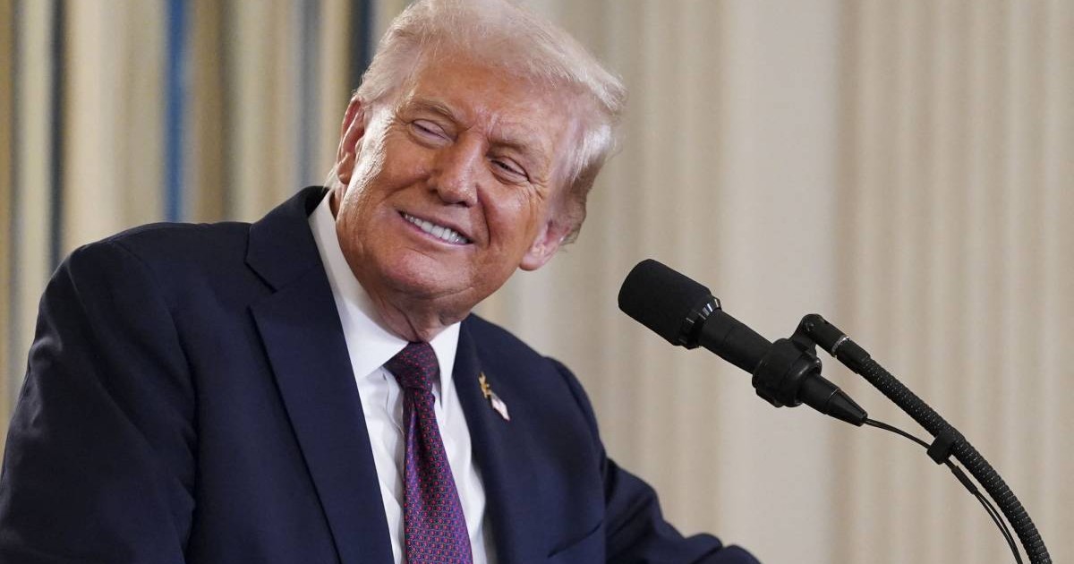 Președintele american Donald Trump, în timpul unei conferințe de presă la Casa Albă, poartă un costum negru și o cravată violet și zâmbește în fața camerei