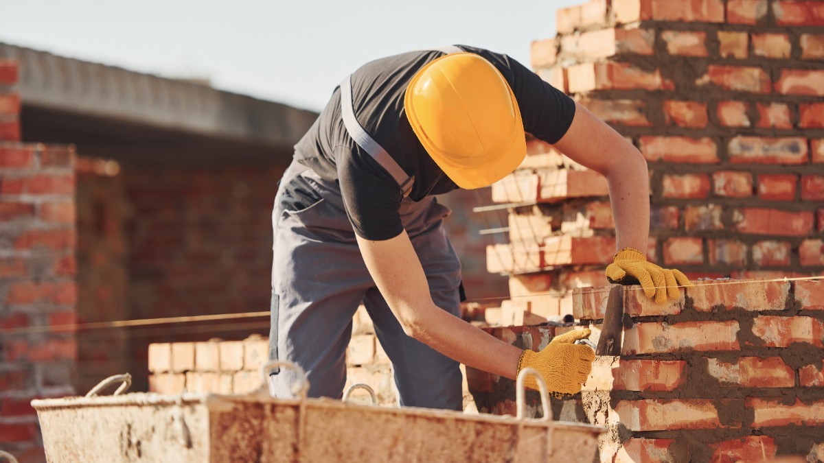 Un muncitor în construcții care poartă cască galbenă de protecție și mănuși, lucrând la zidirea unui perete din cărămidă roșie pe un șantier.