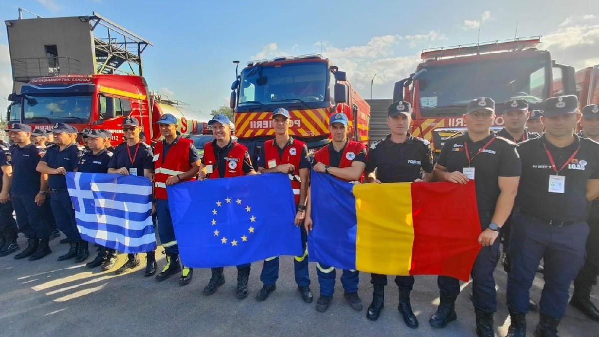 Mai mulți pompieri pozează în fața unor mașini de stingere a incendiilor. ei țin în mână steagul României, al Uniunii Europene și al Greciei
