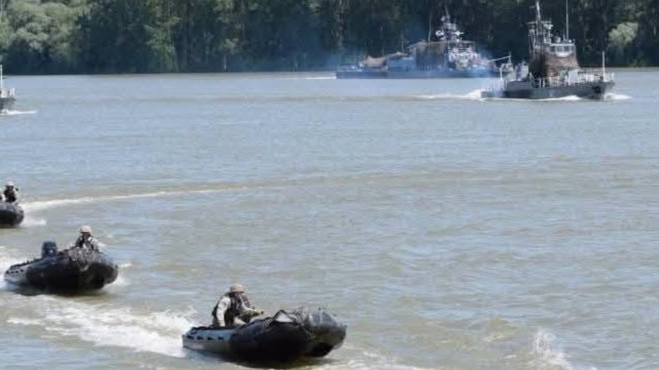 Un exercițiu militar pe apă, în care mai multe bărci pneumatice rapide ghidate de militari înaintează pe un fluviu, având nave militare de patrulare în fundal.