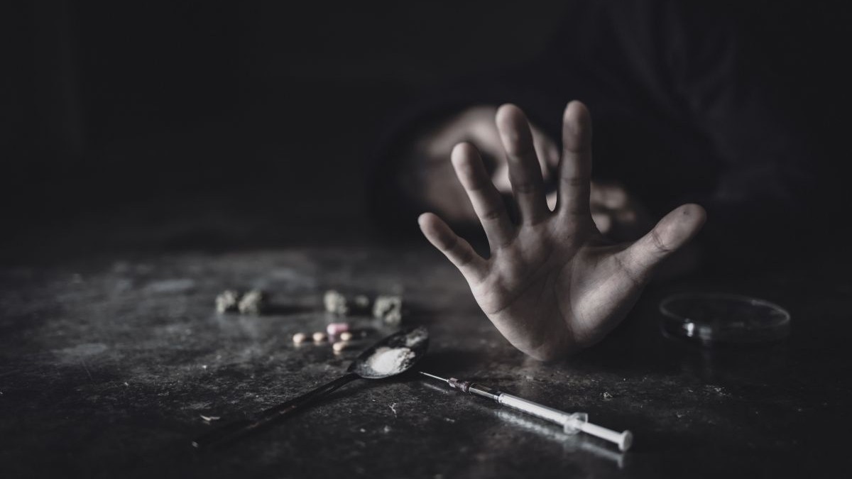 În prim-plan, pe un fond negru, este mâna unei persoane cu degetele deschise, pe masă se vede o seringă și niște droguri, iar în fundal fața persoanei, care stă jos, este acoperită de mână