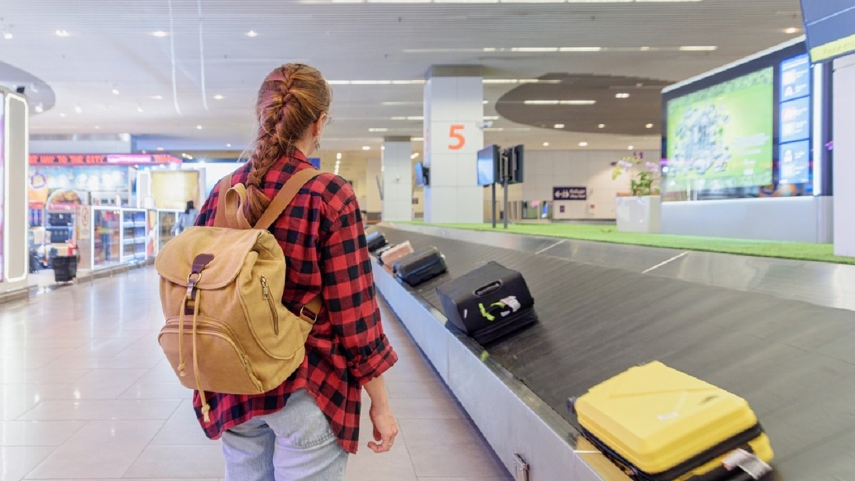 De ce să fotografiezi bagajul înainte de a pleca în vacanță - Imagine cu o femeie cu un rucsac în spate aşteptându-şi bagajul de cală la banda de bagaje dintr-un aeroport