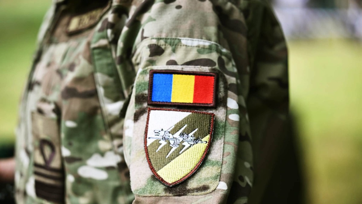 Imaginea prezintă un prim-plan al mânecii unei uniforme militare de camuflaj, pe care este cusut un ecuson cu drapelul României.