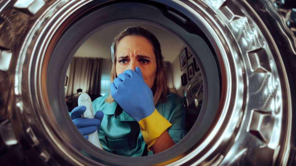 Femeie nemulțumită de o mașină de spălat veche, cu mucegai și miros neplăcut. Poartă mănuși și ține în mână un detergent pentru curățarea aparatului electrocasnic.